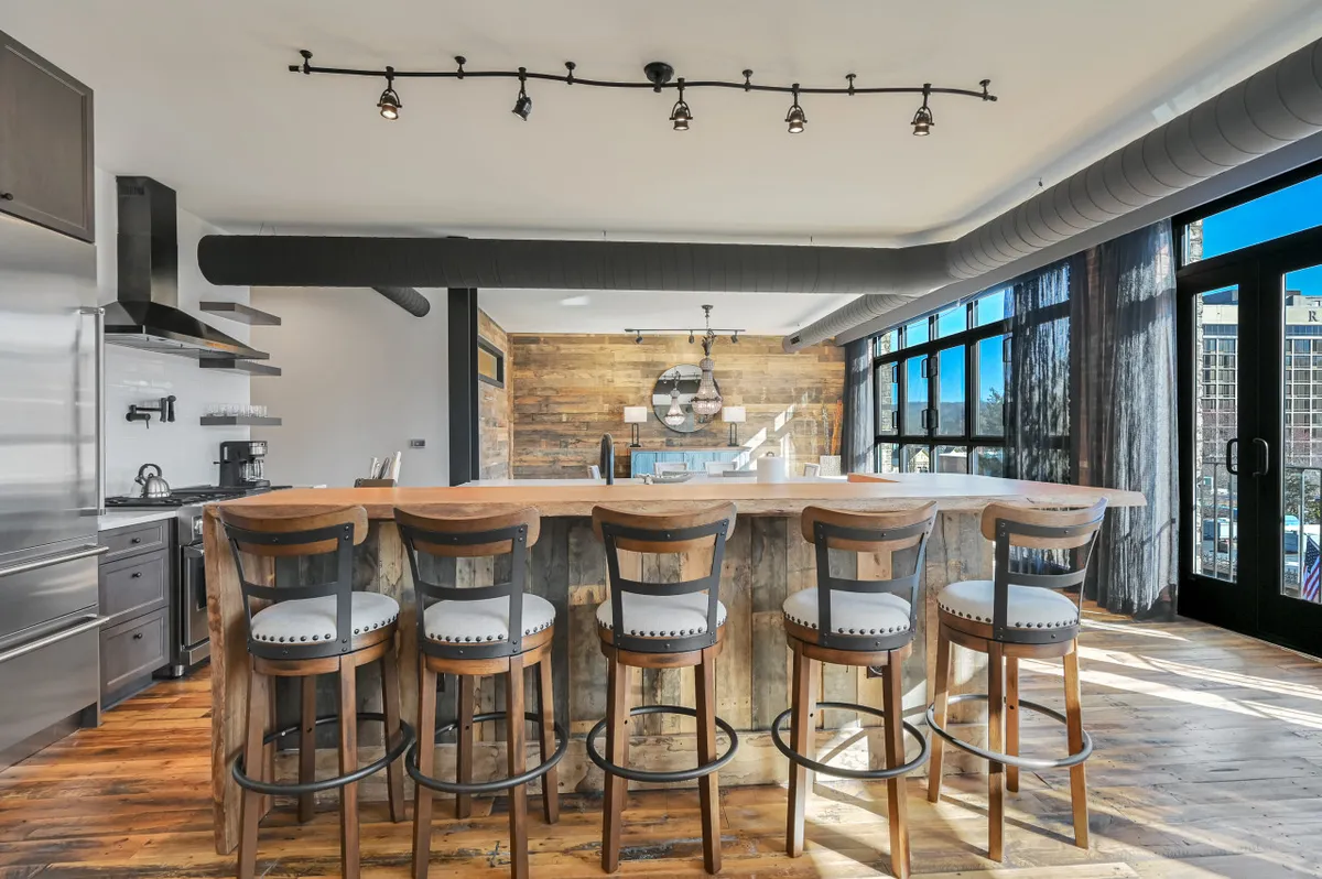 This image focuses on a chic bar counter with stools and ample windows framing a view, accompanied by a wooden accent wall. The arrangement encourages socializing, whether enjoying a cocktail or a casual brunch. The contemporary styling and the window views echo the city-living vibe of the section.