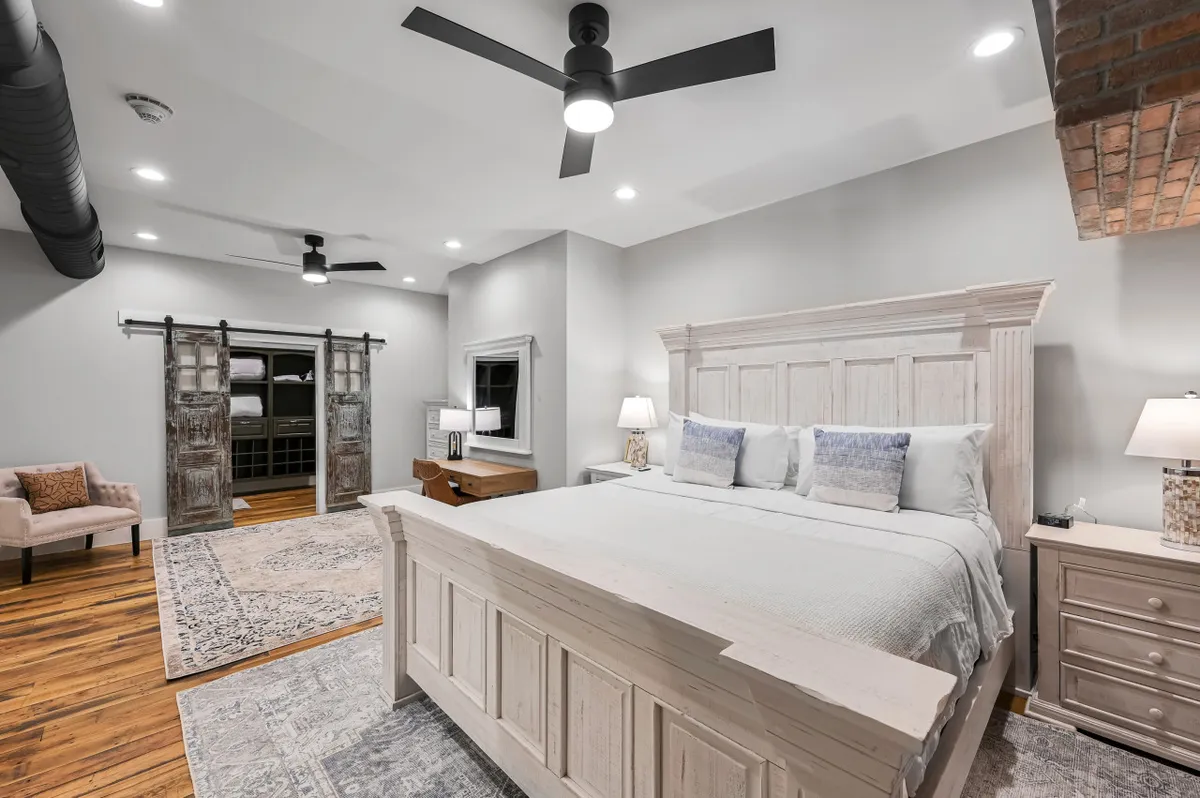 A tranquil bedroom with a king-sized bed, rustic sliding barn door, a decorative chair, and a wooden ceiling fan, creating a warm and inviting retreat.