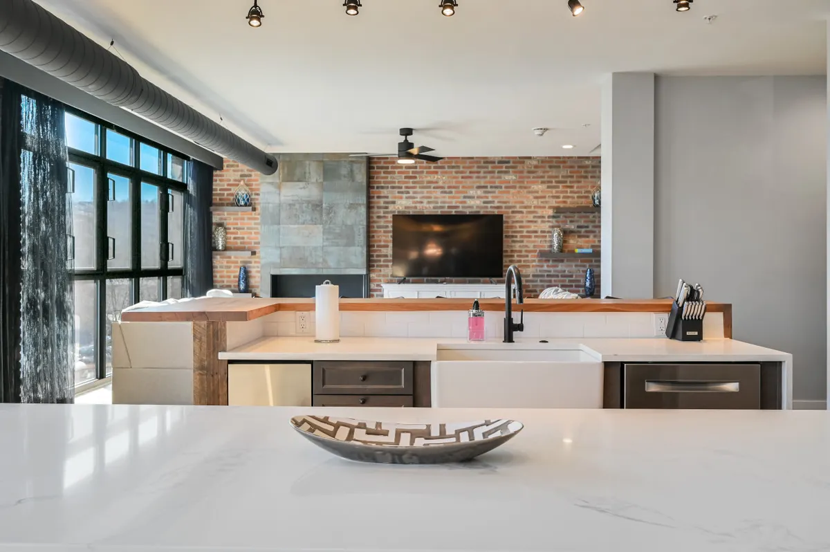 This image captures a welcoming, contemporary open-plan living space featuring a kitchen island, modern sink, exposed brick wall, and a television. The open layout hints at both communal and independent activities, making it ideal for entertaining or unwinding after a workday.