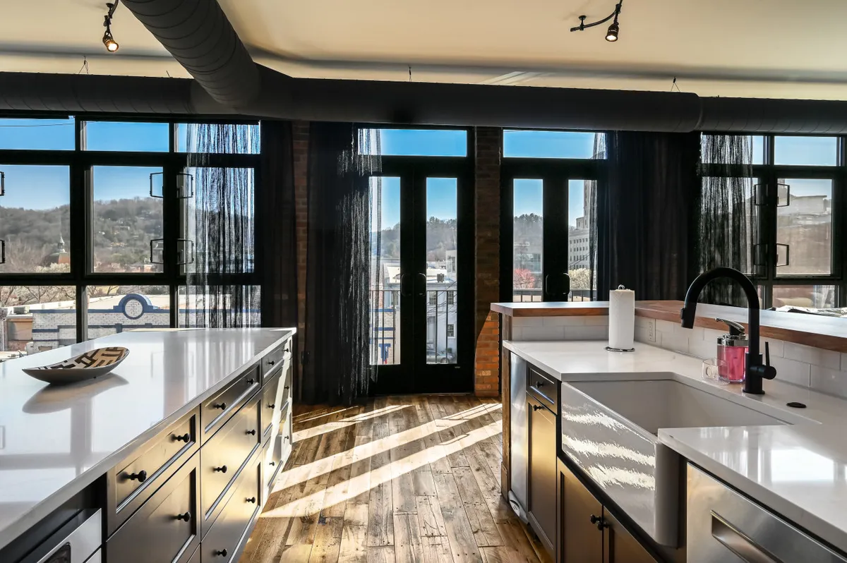 A bright, modern kitchen featuring expansive windows letting in natural light, a central kitchen island perfect for prep or gathering, and sleek countertops. The space is designed for both cooking and entertaining, with contemporary finishes and room for socializing around the island.