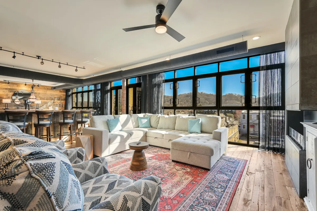 This image highlights a large sectional sofa arranged around a wooden coffee table, placed atop an area rug. The space is defined by expansive floor-to-ceiling windows, allowing abundant natural light to flood the room and offering a clear view of the cityscape beyond. The atmosphere is inviting, with comfortable seating that encourages relaxation and socializing, and the overall openness of the layout emphasizes the room's suitability for gatherings.