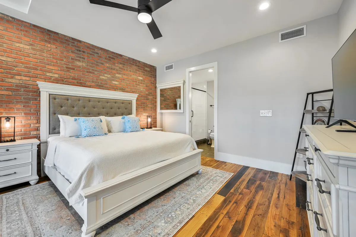 A cozy bedroom with a tufted headboard, exposed brick wall, wooden floor, decorative rug, and a wall-mounted TV. Sunlight filters in, creating a warm environment for unwinding after a productive day.
