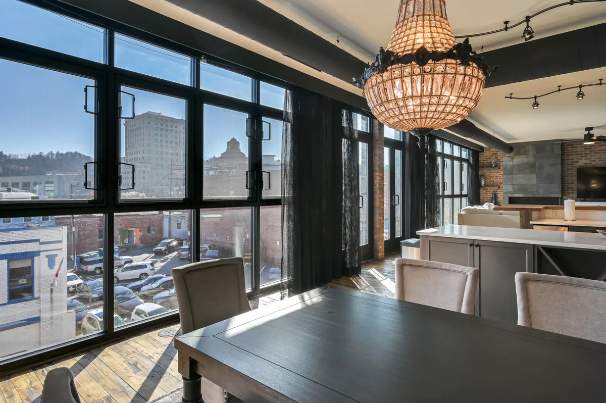 A spacious dining area featuring a large table set near expansive windows, with a modern chandelier overhead. The view through the windows hints at a lively urban environment, suggesting a connection between the interior space and the bustling city outside. The setting exudes warmth and is clearly designed for socializing and group gatherings.