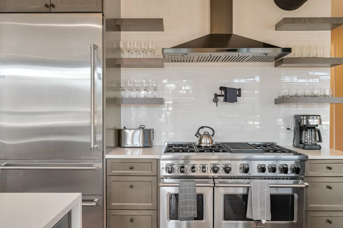 A modern chef’s kitchen with sleek stainless steel appliances, a prominent gas range, and open shelving stocked with glassware. The functional yet stylish setup is ready for cooking and entertaining, evoking the feel of a true culinary workspace.