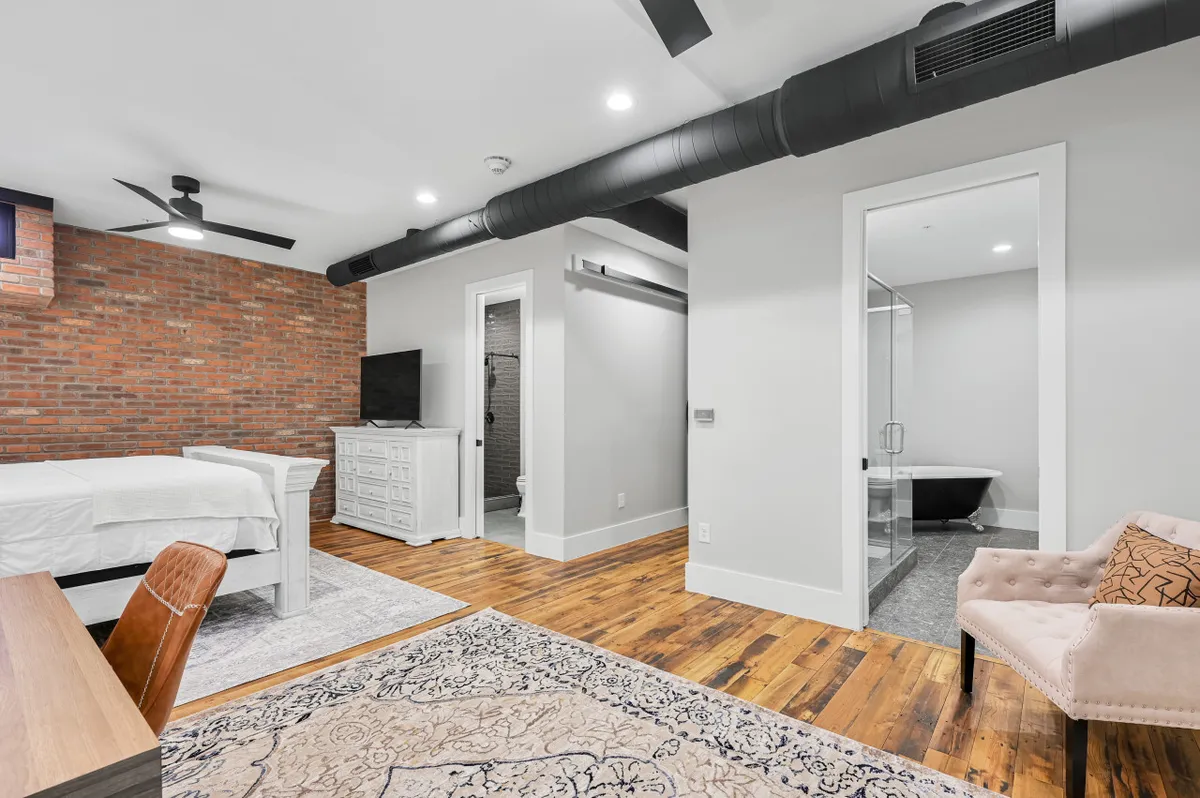 This image showcases an open, stylish space with an exposed brick wall, elegant armchair, work desk, and a glimpse into the bathroom, all set on warm wooden flooring. The atmosphere is both relaxed and energizing, suggesting a comfortable spot to launch or recharge after downtown excursions.