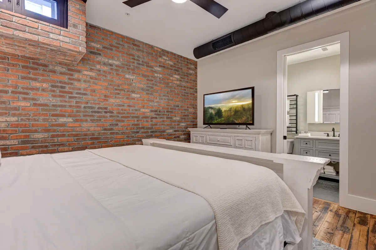 A cozy bedroom with an exposed brick wall, a plush comfortable bed, a large TV, and light wood furniture. The mood is both inviting and restful, suggesting a private retreat after a day exploring downtown.
