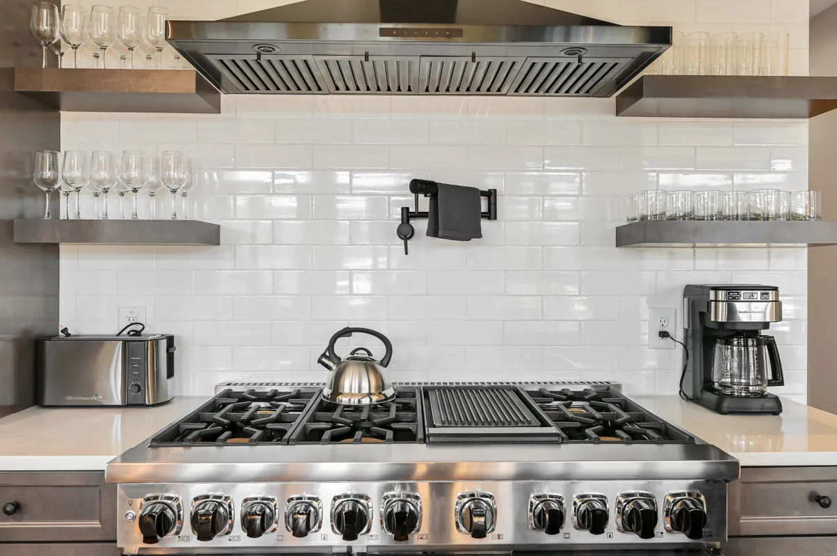 A close-up view of the kitchen’s stainless steel gas range and hood, open shelves with glassware, and a coffee maker, emphasizing the high-end culinary appliances and amenities available for meal prep and hosting brunches.
