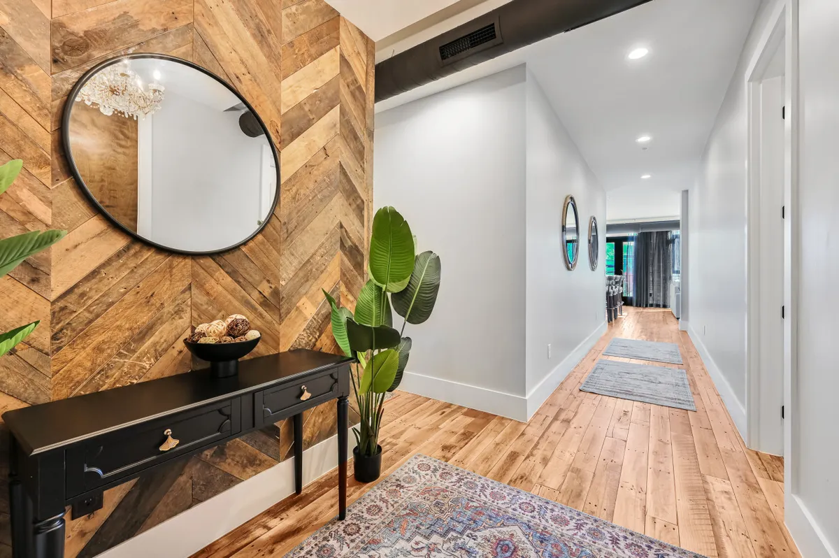 This image shows an inviting entryway featuring a chevron wood wall, a round mirror, a sleek black console table, a green plant, and a decorative bowl. The space is well-lit and thoughtfully styled, evoking a sense of welcome and order.