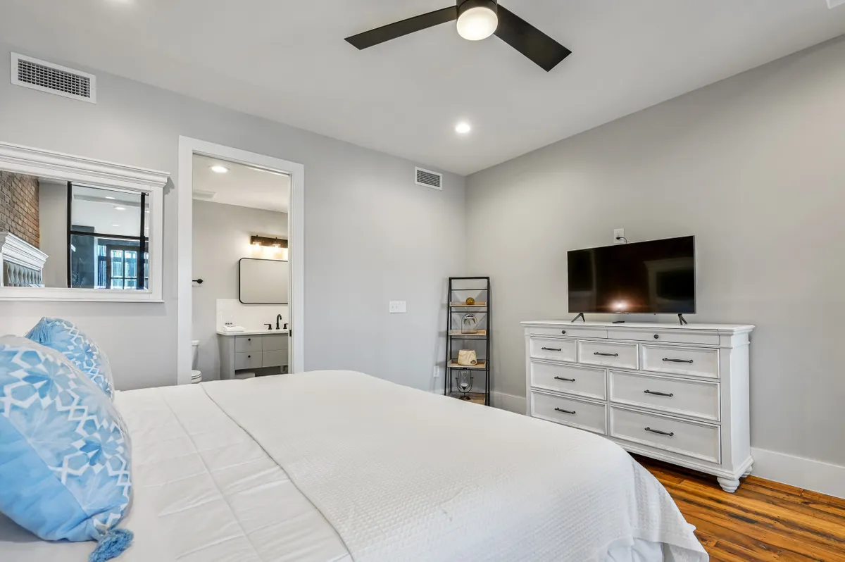 A calm and restful bedroom featuring a plush bed with decorative pillows, a television for entertainment, a dresser for storage, and an open door leading directly to an ensuite bathroom.